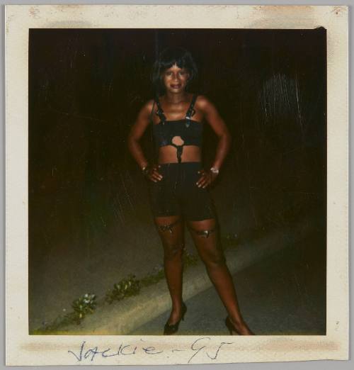 Jackie - 95 [Woman wearing black crop top with harness, short black skirt, and fishnets with her hands on her hips outside at night]