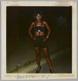 Jackie - 95 [Woman wearing black crop top with harness, short black skirt, and fishnets with her hands on her hips outside at night]