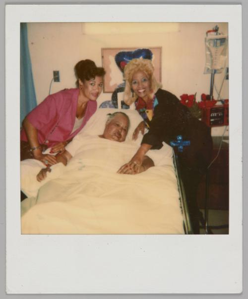 [Man lying down in hospital bed with two women on either side of him, woman on left in pink shirt and woman on right is blonde]