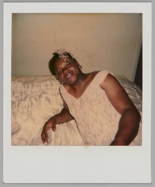 [Person wearing a lace tank top and a pink beaded hairpiece reclining against a bed with shiny sheets]