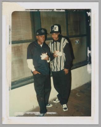 [Twp teenagers posing with one hand in pocket and one hand holding take-out food, person on right wearing white and black striped shirt and "JR" baseball cap]