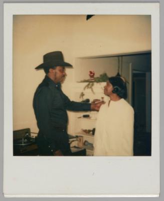 [Man in black shirt and cowboy hat holding a woman dressed in white by her shoulder, they are standing inside]