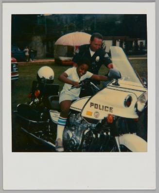 [Boy with white and blue socks sitting on police motorcyce and pretending to drive, officer beside him]