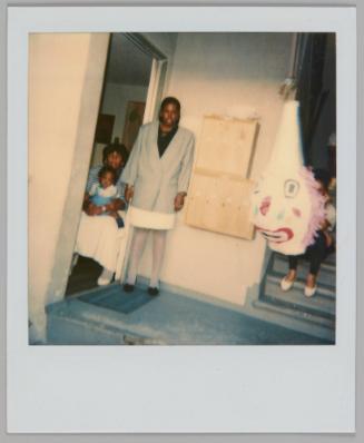 [Woman standing in grey blazer by doorway and beside a woman is sitting with a baby on her lap, large clown head decoration hanging to right]