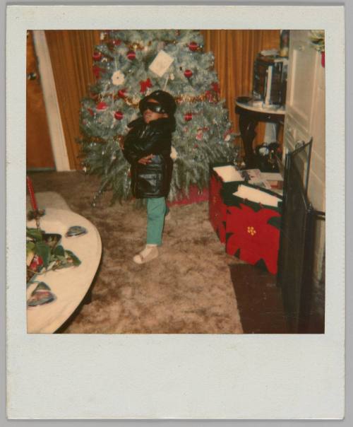 [Boy in black sunglasses, hat and jacket posing with his arms crossed in living room with Christmas tree]