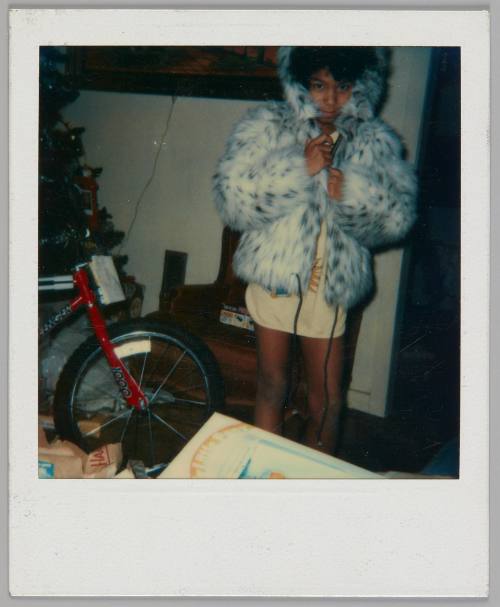 [Young person standing in long t-shirt and wrapping themselves in faux fur coat with the hood up, there is a red bike to the left]