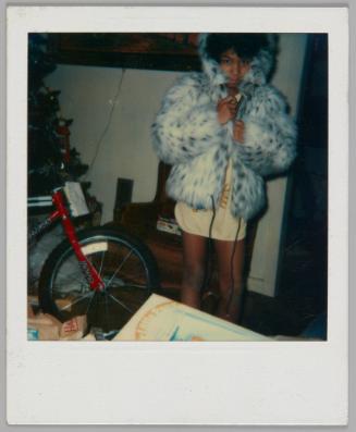 [Young person standing in long t-shirt and wrapping themselves in faux fur coat with the hood up, there is a red bike to the left]