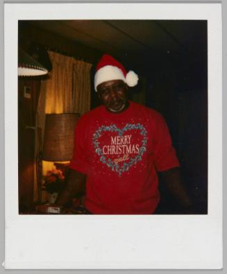 [Man in Santa Claus hat and red sweater with a heart-shaped wreath and the words "Merry Christmas y'all"]