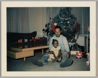 [Girl sitting on the floor between a man's legs with her doll, man is looking down at her, there is a Christmas tree behind them]