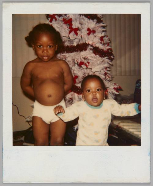 [Young child in underwear with their hands behind their back standing beside a baby with their arms raised, white Christmas tree with red bows behind them]