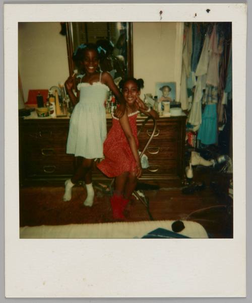 [Two girls, one on left wearing blue dress and one on right wearing red dress, standing in front of dresser beside a closet filled with clothes]