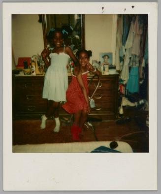 [Two girls, one on left wearing blue dress and one on right wearing red dress, standing in front of dresser beside a closet filled with clothes]