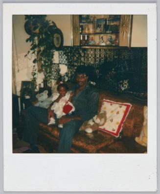 [Man sitting on couch with toddler girl sitting in his lap, stuffed animal beside him]