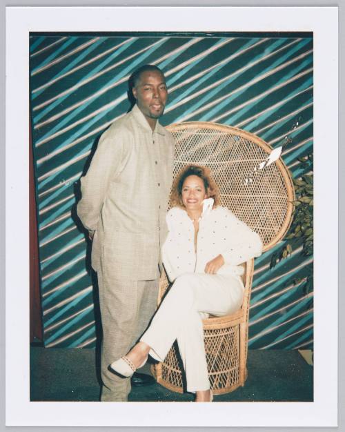 [Young man in light brown outfit standing next to woman in white outfit sitting on larger wicker chair]