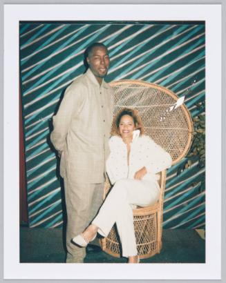[Young man in light brown outfit standing next to woman in white outfit sitting on larger wicker chair]
