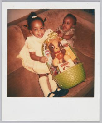 [Infant and toddler girl sitting in corner of couch holding gift basket]