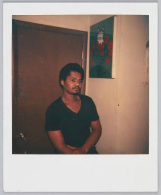 [Man in black vneck tshirt standing in front of door, painting of cat hung on wall behind him]