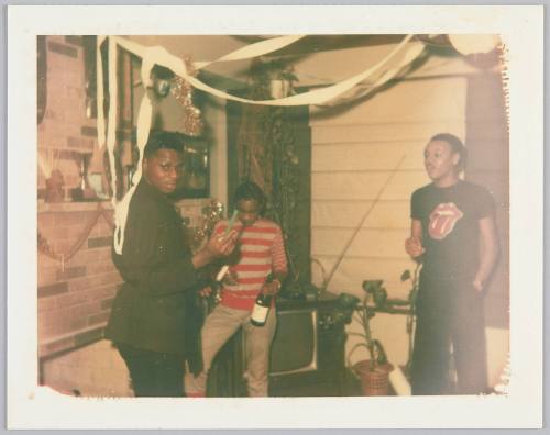 [Three young men standing in corner of room, young man in striped shirt sits on TV set]