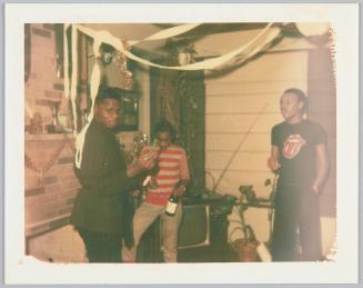 [Three young men standing in corner of room, young man in striped shirt sits on TV set]