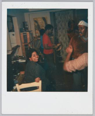 [Woman sitting on chair smiling back at camera, two individuals dancing right of her]
