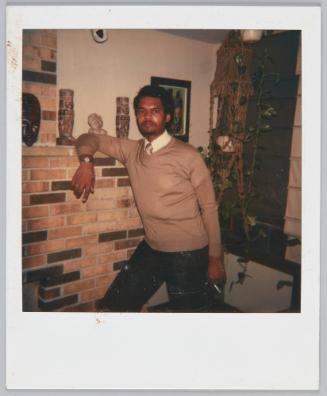 [Man in brown sweater leaning against mantel with a cigarette in hand]