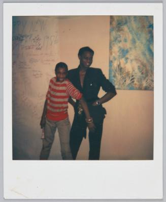 [Two young individuals posing in front of wall, sign in background reads 'Happy New Year 1982']