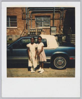 [Two little girls in formal dresses posing in front of car, brick building behind them]