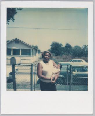 [Woman holding infant outside of house]