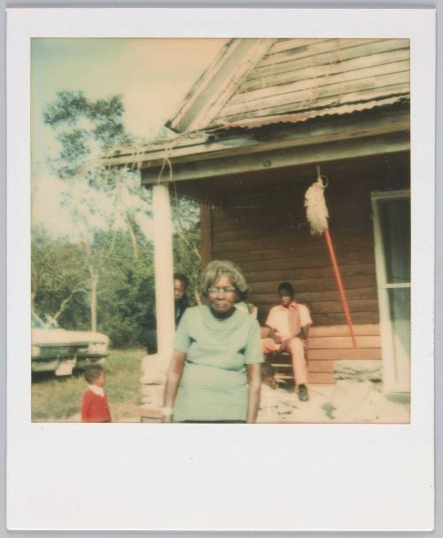 [Elderly woman in blue striped tshirt slightly hunched over, house in the background]