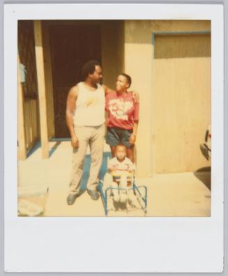 [Young couple with arm around each other standing behind baby sitting on rocking horse]