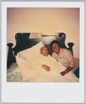 [Woman with glasses posing next to elderly man laying in bed]