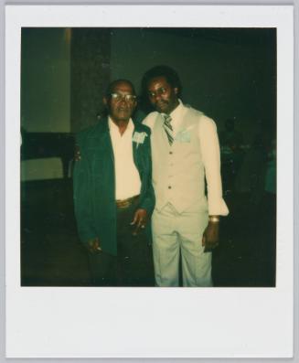 [Two men in formal attired posing, man on right wraps arms around older man]