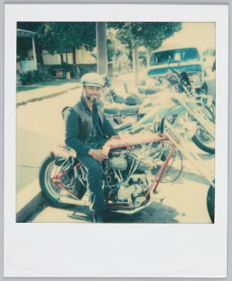 [Man wearing vest and hat smiling while sitting on motorcycle]
