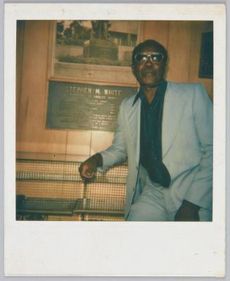 [Older man in grey blazer and pants leaning against bench, behind him reads a sign that says "Stephen M. White"]