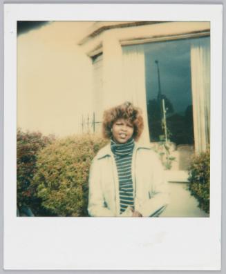 [Young woman wearing striped turtleneck and jacket standing in front of house window]