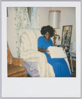 [Elderly woman sitting in armchair, holding and looking at white box in lap]