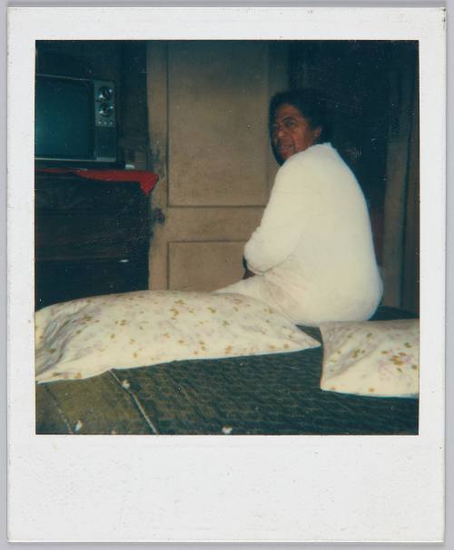 [Elderly woman sitting on edge of bed, two pillows with floral patterns next to her]