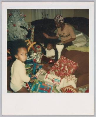 [Toddler and infant surrounded by Christmas gifts, young woman sitting on couch behind them]