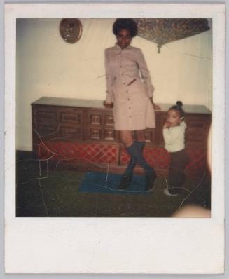 [Young woman posing against cabinet, toddler stands next to her]