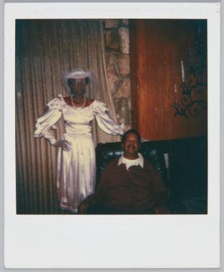 [Woman in pink satin dress and matching hat standing next to older man in recliner chair]