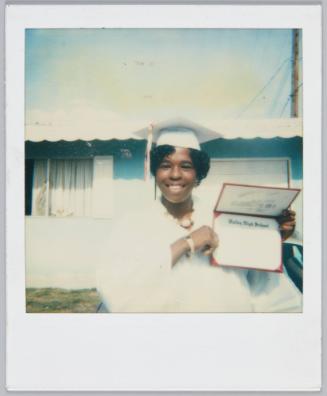 [Adolescent girl wearing white graduation cap and gown smiles while holding up high school diploma]