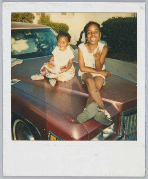 [Little girl and toddler sitting on hood of car, girl making a silly face]