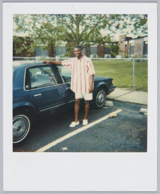 [Young man in white and red vertical striped shirt, and white shorts stands in parking lot with hang on car roof]