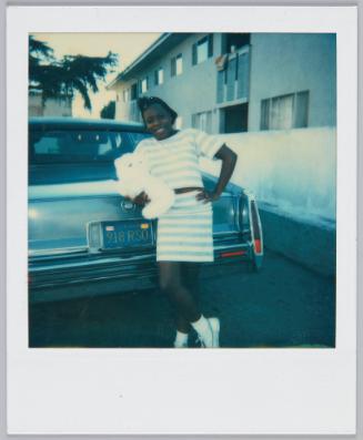 [Girl wearing blue and white stiped top and matching skirt leans against car and holding a white stuffer animal]