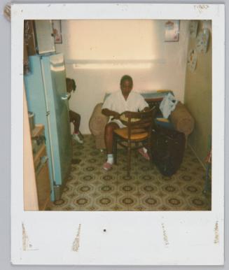 [Woman sitting on seat in kitchen holding a bowl, little girl peeking out behind refrigerator]