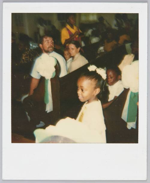 [Little girl in white and matching white hair accessories stands in aisle, people sitting in pews in the background]