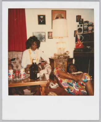 [Woman sitting on couch looking at polaroid photos, individual laying on ground next to her grabbing ankle]