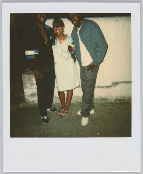 [Woman standing in between two men, all standing in front of white wall]