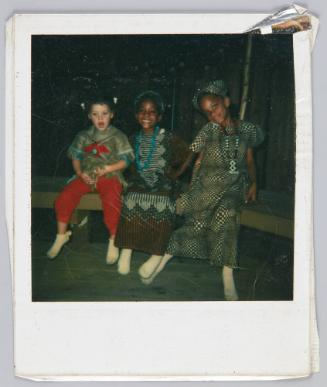 [Three little girls in patterned dresses sitting on bench]