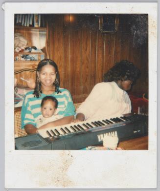 [Two woman sitting in front of YAMAHA keyboard, woman on left holding infant on her lap]
